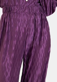 Fioletowe, plisowane spodnie z błyszczącej tkaniny. Posiadają teksturowany wzór fal o elastycznym pasie dla komfortu.