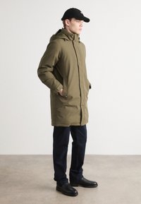 Olivgrüne Parka mit Reißverschluss, Kapuze und seitlichen Taschen, kombiniert mit marineblauen Hosen und schwarzen Stiefeln in einem minimalistischen Umfeld.