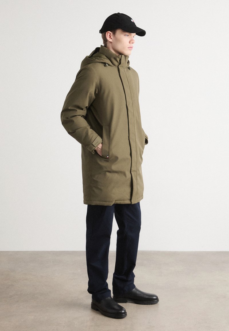 Olivgrüne Parka mit Reißverschluss, Kapuze und seitlichen Taschen, kombiniert mit marineblauen Hosen und schwarzen Stiefeln in einem minimalistischen Umfeld.