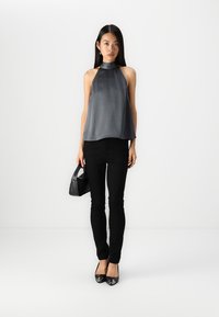 Haut halter gris foncé avec texture plissée, associé à un jean skinny noir et des ballerines noires brillantes. Petit sac à main noir tenu dans la main gauche.