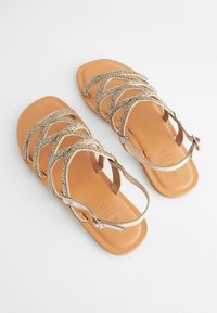 Sandali beige con suola piatta e punta quadrata, caratterizzati da cinturini metallici incrociati. La parte superiore ha una texture intrecciata con una finitura liscia.