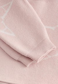 Svetlo ružový sveter s jemnou textúrou. Má rebrované manžety a nenápadný biely geometrický vzor. Materiál vyzerá príjemne a teplo.