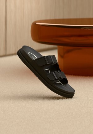 Sort sandal med to stropper og justerbare spænder balanceret på beige tæppe nær en rund, blank bordfod i ravfarve.