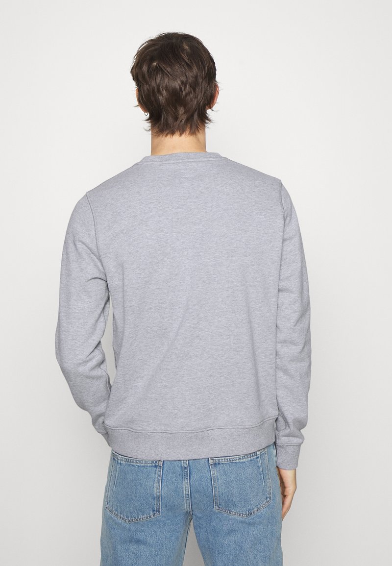 Homme aux cheveux bruns mi-longs portant un sweat-shirt gris clair et un jean bleu, debout de dos devant un fond uni.