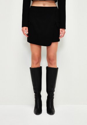 Femme portant une mini-jupe noire et des bottes en cuir noires montant jusqu'aux genoux, debout sur un fond clair uni.