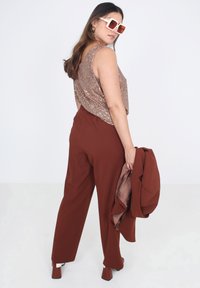 Bruine gepaste broek gecombineerd met een bronzen top met pailletten. Het model houdt een bijpassende bruine blazer vast. Kenmerken zijn oversized beige zonnebrillen en hoger-schoen.