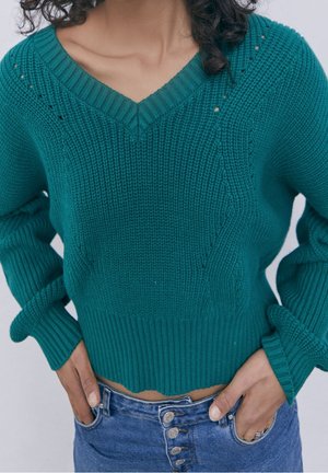 Kobieta ubrana w turkusowy, dziergany sweter z dekoltem w serek i długimi rękawami, w połączeniu z wysokim stanem niebieskich dżinsów, z rękami opartymi na biodrach.