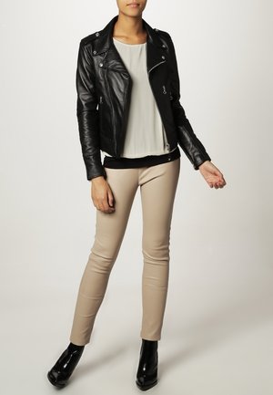 Femme portant une veste en cuir noire, un pantalon beige, un chemisier blanc et des bottines noires, debout avec un bras légèrement tendu.