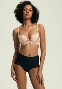 Frau mit lockigem Haar, die einen nudefarbenen Spitzen-BH und hochgeschnittene schwarze Unterhosen trägt, vor einem schlichten hellen Hintergrund.