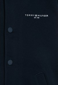 Námornícky modrý sveter s dvoma gombíkmi, s bielym logom "TOMMY HILFIGER" a textom "EST. 1985". Mäkká tkanina s hladkou textúrou.
