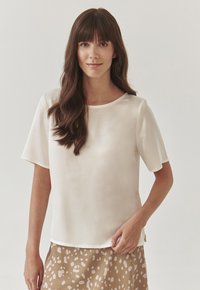 Blusa corta de manga corta color beige claro con una textura suave, escote redondo y un ajuste relajado, combinada con una falda marrón con manchas.