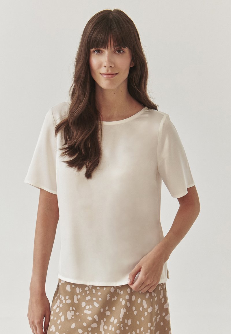 Blusa corta de manga corta color beige claro con una textura suave, escote redondo y un ajuste relajado, combinada con una falda marrón con manchas.