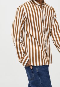 Homme portant une chemise à boutons à rayures verticales marron et blanches avec une poche poitrine, associée à un jean en denim bleu, main dans la poche.