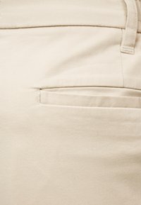 Gros plan sur le tissu beige d'un pantalon montrant une poche passepoilée arrière avec des coutures détaillées et une passants de ceinture au-dessus.