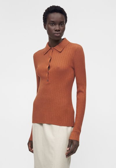 Γυναίκα που φοράει μακρυμάνικη ριγωτή μπλούζα πόλο σε σκουρό πορτοκαλί χρώμα και ανοιχτό μπεζ φούστα, στέκεται μπροστά σε απλό φόντο.