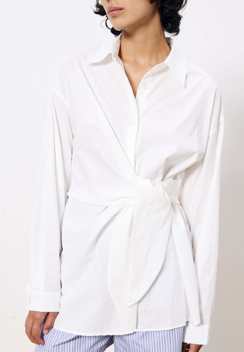 Chemise en coton blanche à manches longues, avec un devant boutonné et un détail de taille nouée. Texture lisse, coupe décontractée, assortie à un bas à rayures bleues.