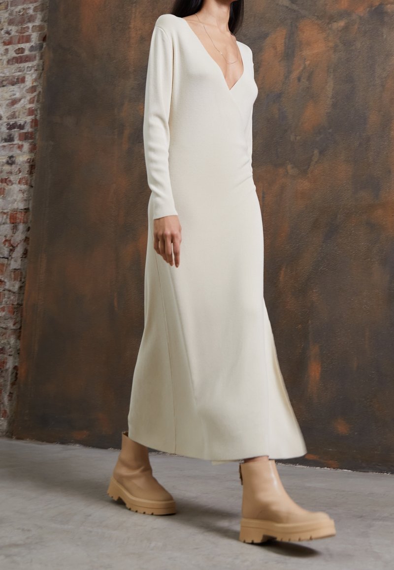 Robe longue en maille de couleur crème avec un décolleté en V profond et des manches longues, présentant une coupe ajustée et une fente latérale. Assortie à des bottes beige à semelle épaisse.