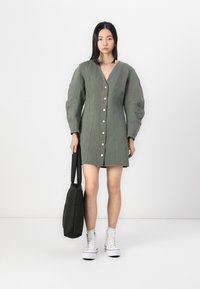 Zelena denim obleka z V-izrezom, gumbi spredaj in napihnjenimi dolgimi rokavi. Model drži črno torbico, kombinirano z belimi visokimi supergami.