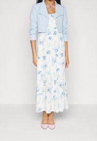Longue robe blanche à motif floral bleu, jupe à volants, associée à une veste courte bleu clair. Chaussures pointues roses complètent la tenue.