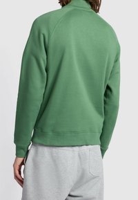 Grüner Pullover mit hohem Kragen, gerippten Bündchen und Saum. Verfügt über eine glatte Textur und Raglanärmel. Kombiniert mit grauer Jogginghose.
