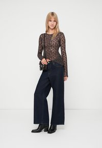 Top de encaje de manga larga en marrón oscuro, combinado con jeans de mezclilla oscura de pierna ancha. La modelo lleva botas negras hasta el tobillo y sostiene un pequeño bolso negro.