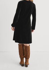 Svart sammet långärmad klänning med A-linjeform och rundad halsringning, ihop med knähöga bruna mockastövlar.