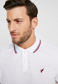 Polo bianco con un placket a due bottoni, colletto a strisce rosse e blu in contrasto, e un piccolo logo ricamato rosso sul petto.