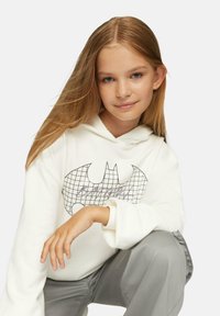 Biała bluza z kapturem z czarnym gridowym logo Batmana, wykonana z miękkiego materiału i o luźnym kroju, zestawiona z szarymi spodniami i widocznymi mankietami.