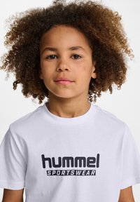 Vit bomull T-shirt med "hummel" och "SPORTSWEAR" tryckta i fet svarta bokstäver över bröstet, korta ärmar och rund halsringning.