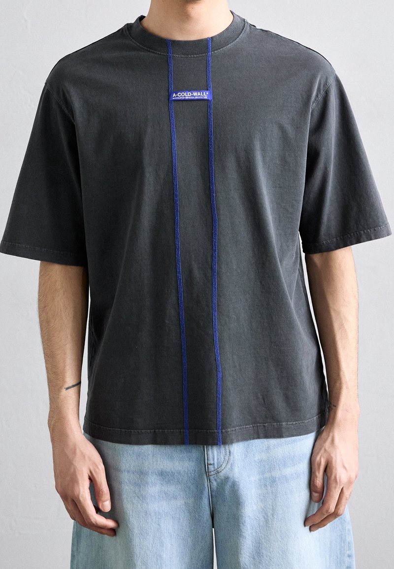 Homme portant un T-shirt noir ample avec des coutures verticales bleues et une étiquette "A-COLD-WALL" sur la poitrine, associé à un jean bleu clair.