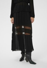 Čierna plisovaná maxi sukňa s priehľadnými panelmi a vrstveným dizajnom, s jemnou textúrou a plynulou siluetou. Kombinovaná s čiernymi členkovými čižmami.