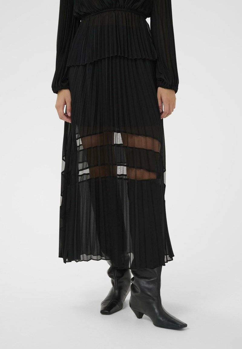 Čierna plisovaná maxi sukňa s priehľadnými panelmi a vrstveným dizajnom, s jemnou textúrou a plynulou siluetou. Kombinovaná s čiernymi členkovými čižmami.