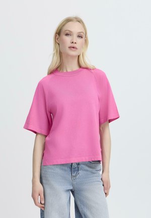 Blonde Frau, die ein leuchtend pinkes Kurzarmoberteil und hellblaue, hochgeschnittene Jeans trägt, steht vor einem schlichten weißen Hintergrund.