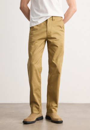 Mężczyzna w beżowych prostych spodniach, beżowych zamszowych butach i białej koszulce, stojący z rękami za plecami na szarej podłodze.