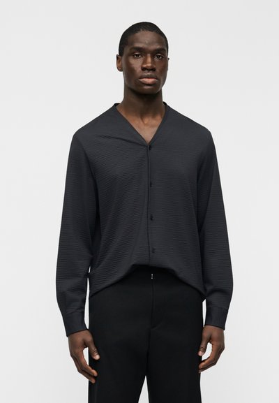 Chemise noire texturée à manches longues avec col en V et boutons à l'avant, présentant une coupe décontractée et des poignets côtelés, portée avec un pantalon noir.