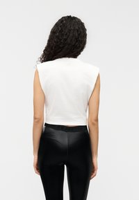Λευκό κοντό τοπ με φαρδιά σχεδίαση στους ώμους. Συνδυάζεται με μαύρο κολάν υψηλής λάμψης με ελαστική μέση. Μινιμαλιστική λεπτομέρεια με λογότυπο.