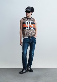 Νέος άνδρας που φοράει αμάνικο τοπ με λεοπάρ εκτύπωση και αριθμό 95, σκούρο τζιν, μαύρο καπέλο, μεγάλα γυαλιά ηλίου και μαύρα αθλητικά παπούτσια, που στέκεται χαλαρά.