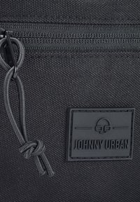 Čierna látková taška s uzatváraním na zips, prešitými detailmi, gumenou nášivkou loga a textúrovaným povrchom.