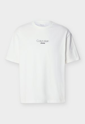 Balta T-krekls ar īsām piedurknēm, uz krūtīm centrēti melniem burtiem "Calvin Klein JEANS" vienkāršā fona.