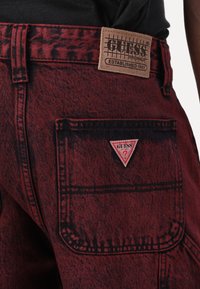 Røde denimshorts med et tekstureret mønster, der har et GUESS-logo-patch og lomme detaljer, metalknap og kontrasterende syninger.