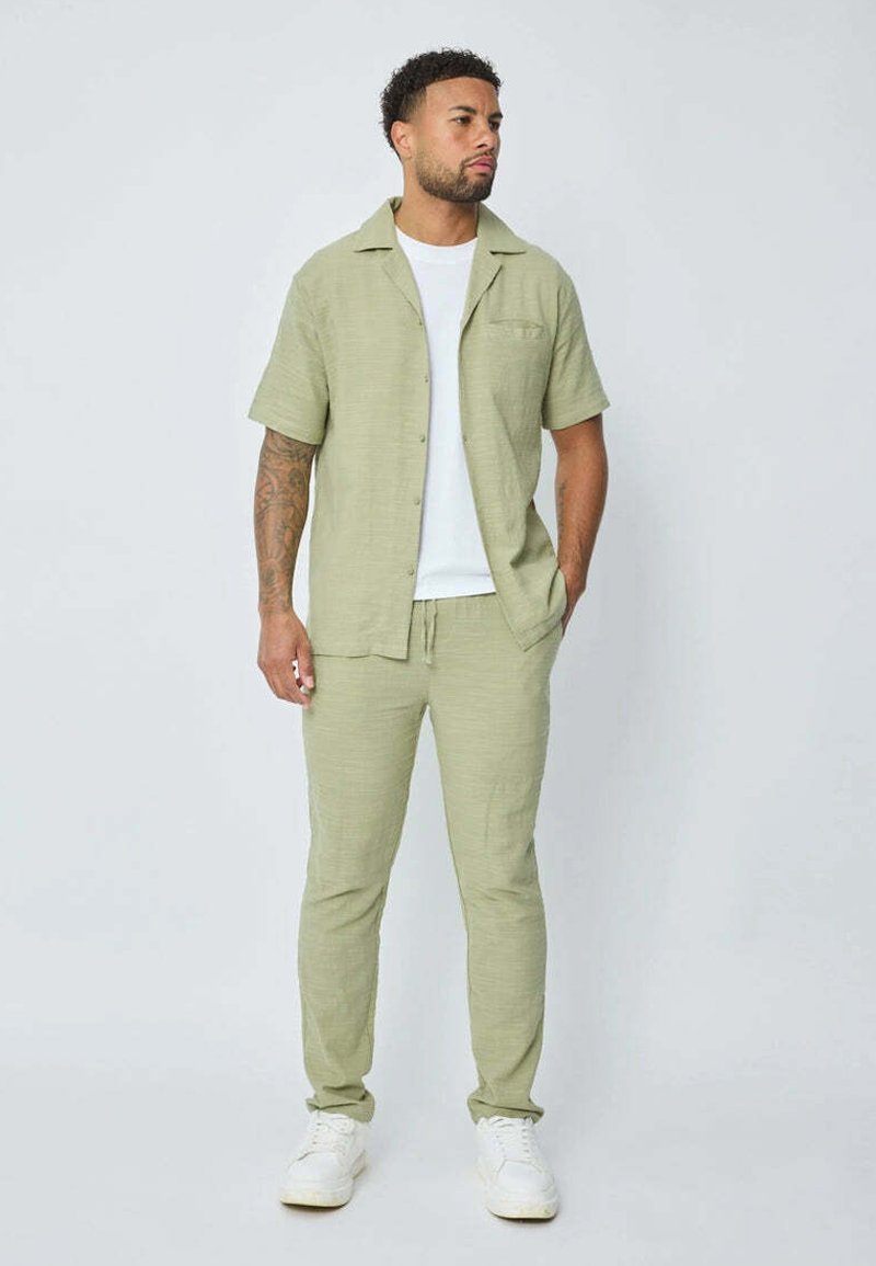 Chemise en lin verte à manches courtes avec une poche, assortie à un pantalon slim-fit. Portée avec un t-shirt blanc et des baskets blanches.