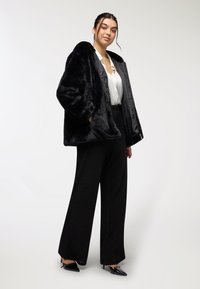 Giacca in pelo sintetico nera con fodera, blusa bianca e pantaloni a gamba larga neri a vita alta. Tacchi neri a punta completano l'outfit.