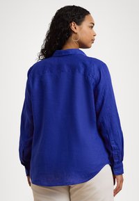 Camisa de linho azul com mangas longas, punhos abotoados, uma pala nas costas e um acabamento inferior curvado. A textura parece leve e respirável.