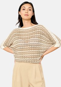 Beige gestrickter Pullover mit lockerer Passform, kurzen Ärmeln, abwechselnden offenen und festen Streifen sowie einem weiten Ausschnitt. Kombiniert mit hellen Hosen.