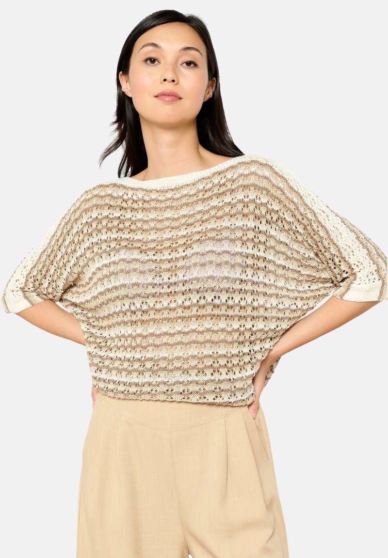 Beige gestrickter Pullover mit lockerer Passform, kurzen Ärmeln, abwechselnden offenen und festen Streifen sowie einem weiten Ausschnitt. Kombiniert mit hellen Hosen.
