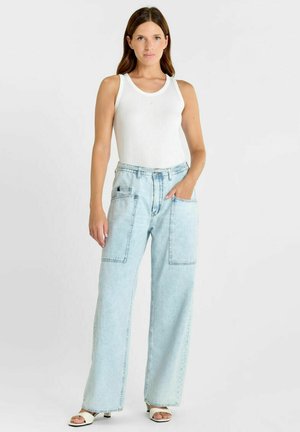 Femme portant un débardeur blanc, un jean taille haute bleu clair à jambes larges avec de grandes poches avant, et des talons blancs ouverts à l'avant.