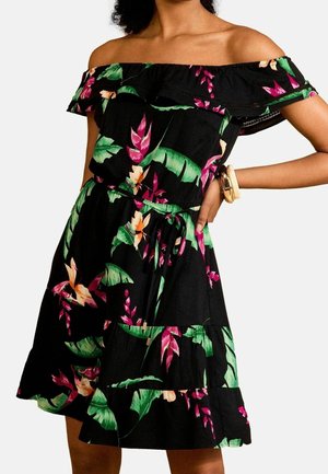 Femme portant une robe noire épaules dénudées avec des motifs floraux tropicaux verts et roses, tenant une main sur sa hanche avec des bracelets au poignet.