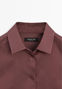 Gros plan sur le col d'une chemise mauve à boutons avec une étiquette "Massimo Dutti" cousue à l'intérieur de l'encolure.