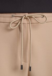 Pantalon beige avec un cordon de serrage noué en nœud décoratif à travers des œillets en métal à la taille, porté avec un haut noir.