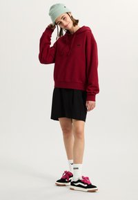 Burgundy hooded sweatshirt, black skirt, black sneakers with pink laces, white socks. Light blue knit beanie; model is posing.